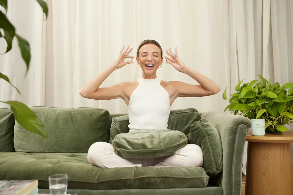 Happy woman sitting cross-legged on sofa, smiling, with hands near her head in a peaceful home setting.